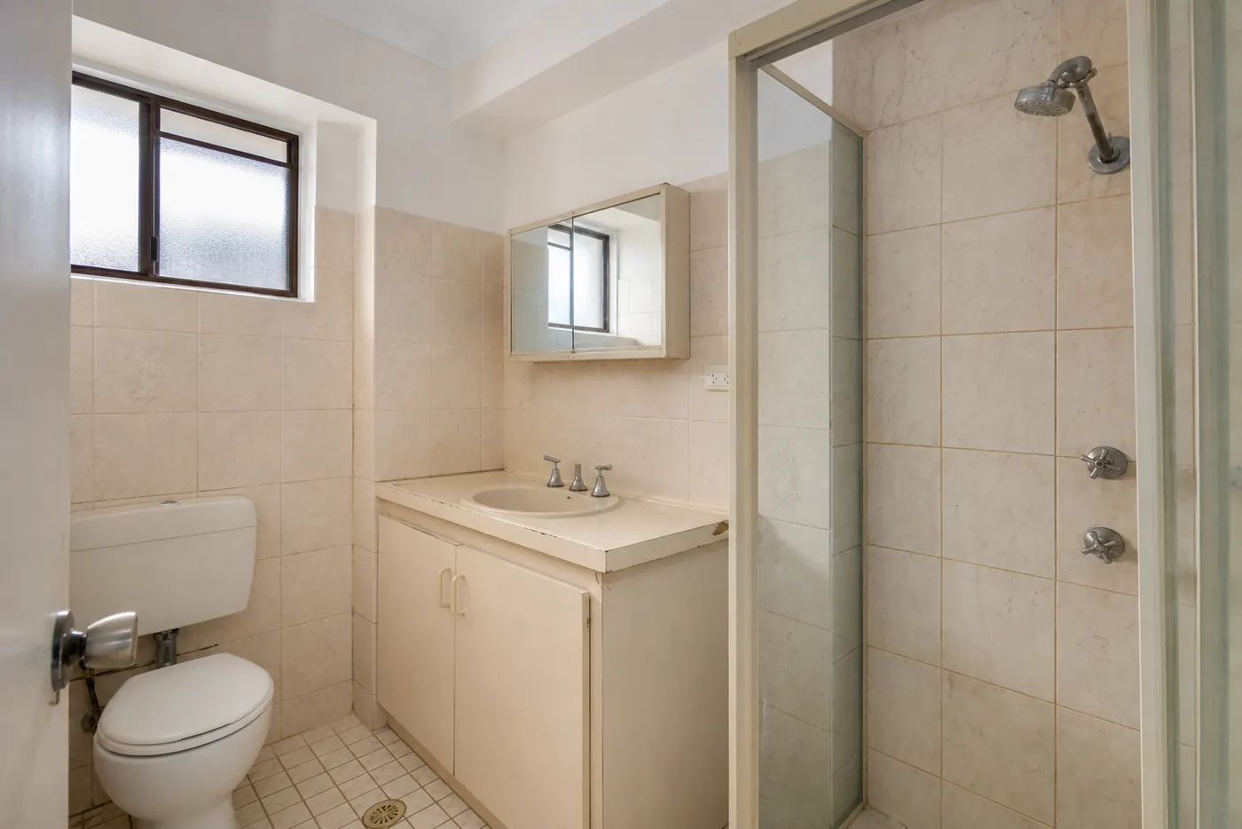 Small ensuite before the renovation: dated cream wall tiles, a chipped laminate vanity top and an old chrome shower head.